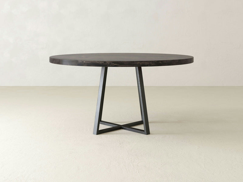 Introducing the Watson Round Dining Table by James+James, featuring a sleek, minimalist design. This round dining table boasts a solid hardwood tabletop with a dark finish and is supported by a striking black, geometric metal base with four legs that converge at the bottom for an industrial look. The backdrop includes a plain, light-colored wall and floor.