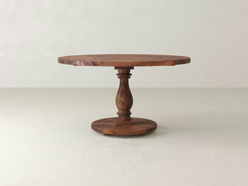 The Vivien Round Dining Table - Tuscany by James+James is a beautifully crafted piece featuring a detailed design with a single intricately carved central pedestal leg that supports the tabletop. This table boasts a flat round base and a smooth wood finish, set against a plain, light-colored background.