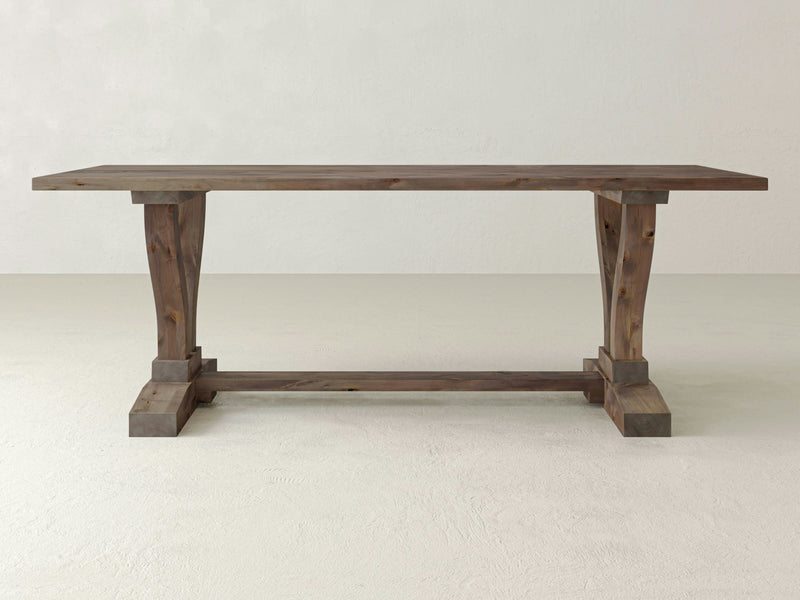 A Vera Dining Table - Barn Wood by James+James, featuring a rustic design, is centered in a minimalist, light-colored room. The rectangular table has a thick top and sturdy, uniquely shaped legs with a horizontal support beam at the base. The natural finish of the wood highlights visible grain patterns.