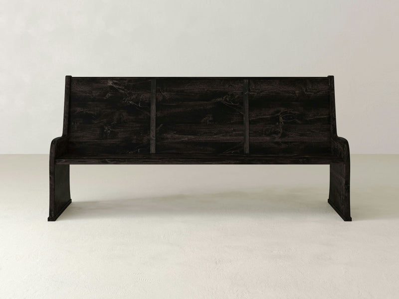 The Parish Pew Bench - Charred Ember by James+James is a dark wooden bench featuring a straightforward, rectangular design and a backrest evenly divided into three sections. It stands on a light-colored floor and comes without armrests or cushions. The background is a plain, light-colored wall.