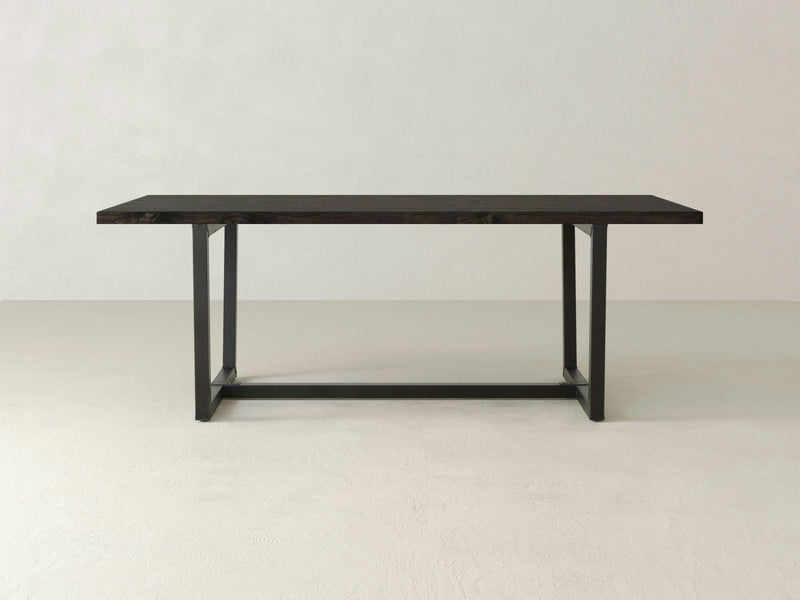 The Trapezoid Dining Table - Charred Ember by James+James features a dark rectangular wooden top and sturdy black metal frame legs. Positioned on a light-colored floor against a plain, light grey wall, this minimalist piece boasts a simple yet elegant design that complements various interior styles.