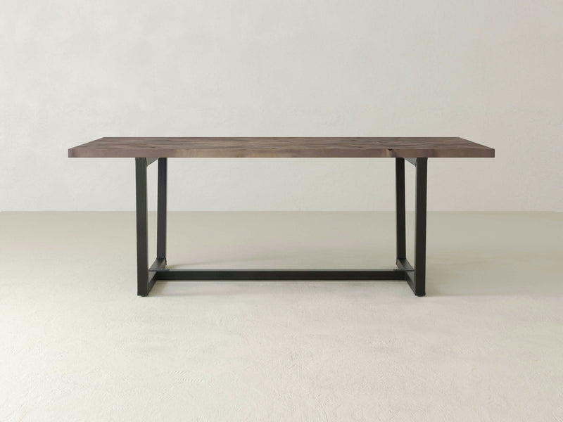 A James+James Trapezoid Dining Table - Barn Wood is set against a plain, light-colored background. The minimalist wooden table features a dark, rectangular top and black metal frame legs in a modern and clean design, complete with a sturdy base connecting the legs.