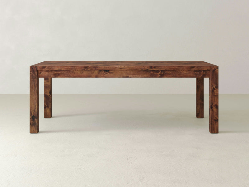 A spacious, wooden rectangular Jonathan Dining Table by James+James with a simple farmhouse style design, featuring thick, straight legs and a smoothly dark-stained surface. The hardwood table is situated in an empty, light-colored room with minimal decoration, highlighting its stark, rustic charm.