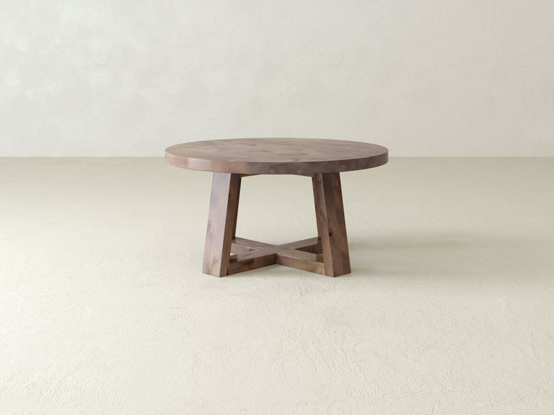 The Rae Coffee Table by James+James, featuring a circular shape and thick top, serves as a centerpiece on a neutral-colored floor with a plain, light backdrop. Its solid, intersecting legs complement the minimalist, rustic aesthetic.