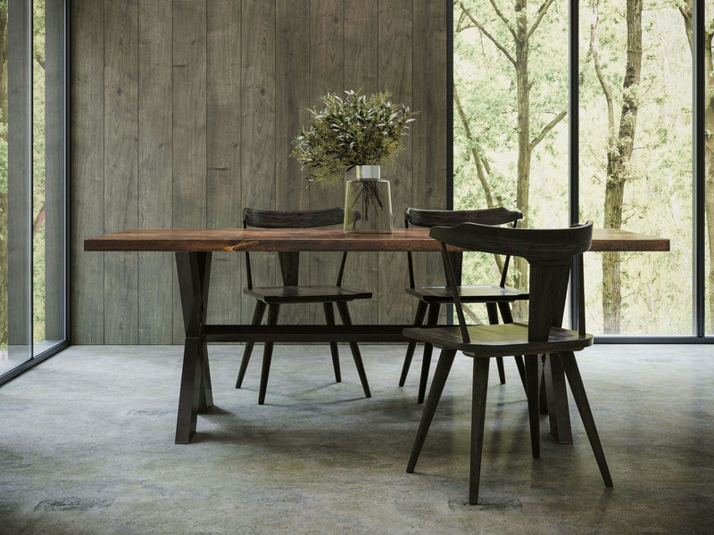 A minimalist dining room featuring the X-Base Dining Table - Tobacco by James+James, paired with four black chairs. The setup is positioned in front of floor-to-ceiling windows and a wooden-paneled wall. At the center of the table, there's a vase filled with greenery, complementing the serene view of a forested area outside.