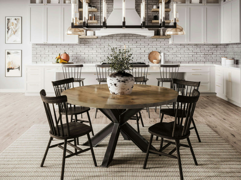 A contemporary dining area showcases the Shiloh Round Dining Table - Harvest Wheat from James+James, adorned with a potted plant centerpiece. Surrounding the table are six black spindle-back chairs. The background features a kitchen equipped with white cabinetry, a light brick backsplash, and illuminated by three pendant lights.