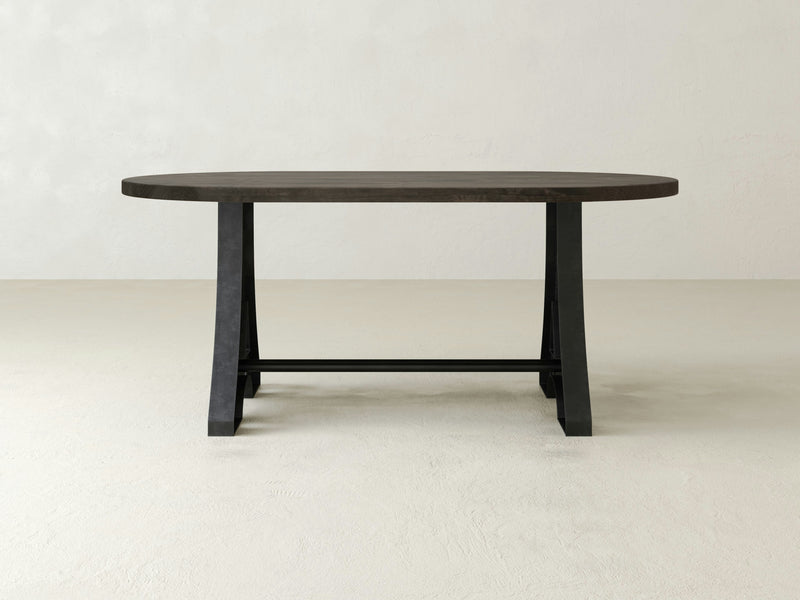 The Wishbone Oval Dining Table - Deep Grey by James+James, with its minimalist design and dark finish, sits on a pale, textured surface. The table features a sturdy base with two legs connected by a horizontal support bar. The background is plain and light-colored, creating a simple and modern aesthetic.