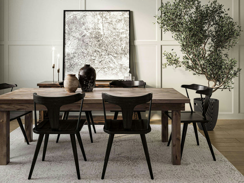 A dining room with a rustic James+James Farmhouse Dining Table - Barn Wood and black chairs. A large abstract art piece in a black frame leans against the paneled wall. A potted olive tree stands in the corner, and various ceramic vases and candlesticks decorate the tabletop.