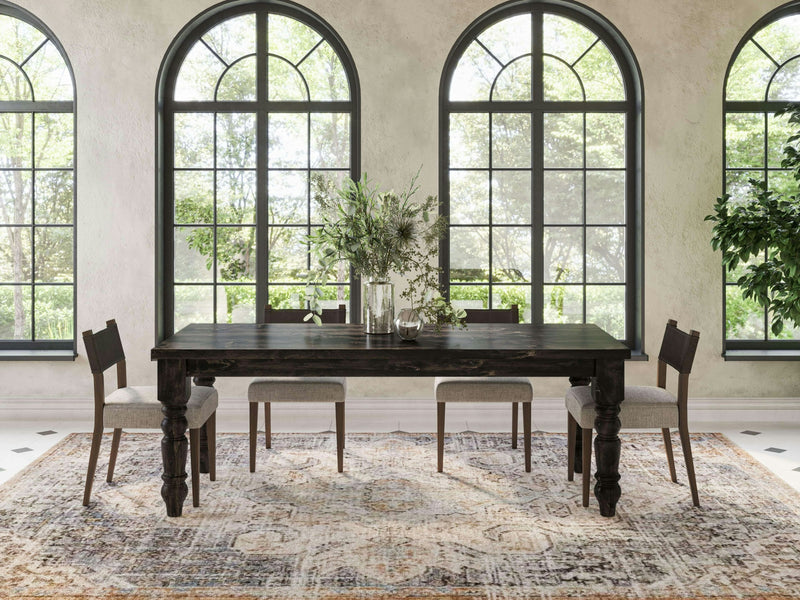 A dining room featuring the James+James Baluster Dining Table in Charred Ember, complemented by four chairs. The table is elegantly adorned with vases of greenery. Large arched windows flood the room with natural light, and a patterned rug covers the floor. The space is decorated with potted plants.