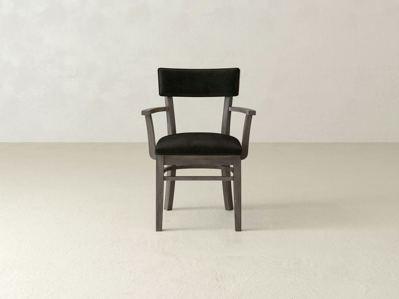 A sleek, modern Everly Arm Dining Chair in deep grey by James+James is placed against a plain, light-colored background. The chair features a cushioned seat and backrest, with wooden legs and a minimalist design.