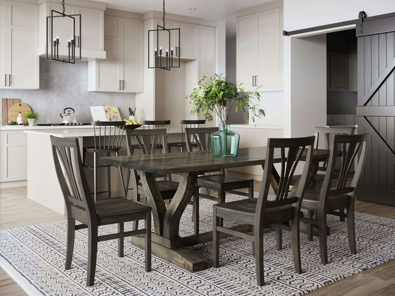 Modern dining room featuring a Vera Dining Table in Deep Grey by James+James and six matching chairs on a patterned rug. Behind the table, there is an island with three black chairs, white cabinets, and a grey backsplash. A sliding barn door and a large green vase with foliage on the table enhance the decor.