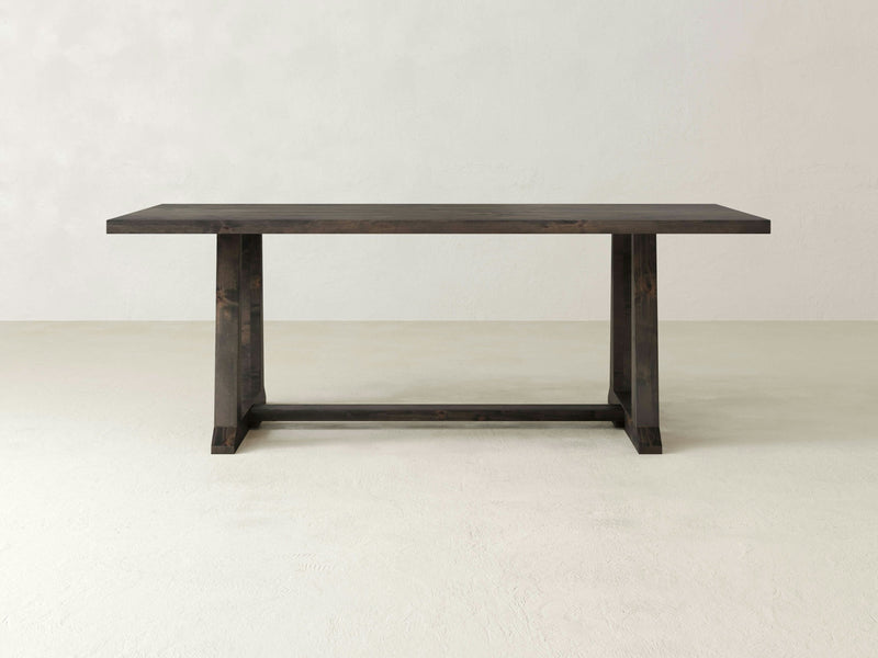 The Cora Dining Table - Deep Grey by James+James features a modern design with a dark wood, rectangular top and sturdy, straight legs that form a T-shape at each end. It is elegantly placed in a minimalist setting with light-colored flooring and wall background.