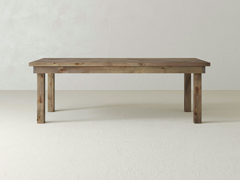 The Farmhouse Dining Table - Barn Wood by James+James stands in an empty room with light-colored walls and floor. The table features a simple, rustic design with a flat rectangular top and four sturdy legs, all made from the same wood. Its natural finish beautifully highlights the wood grain.