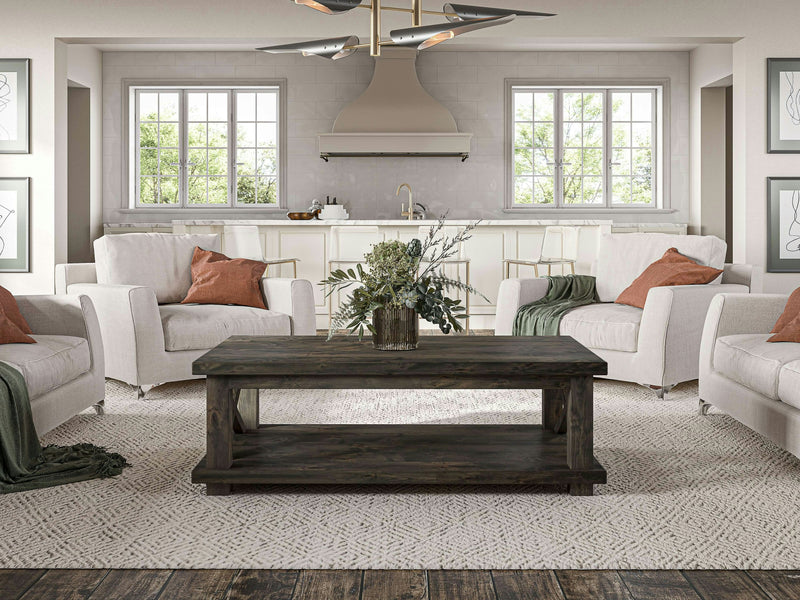 A cozy living room with beige armchairs and white cushions, accented by green and rust-colored throw pillows. At the center is a James+James Carter X Coffee Table in Deep Grey, adorned with a vase of greenery. Behind it, there's a white kitchen featuring a stainless steel hood and large windows.