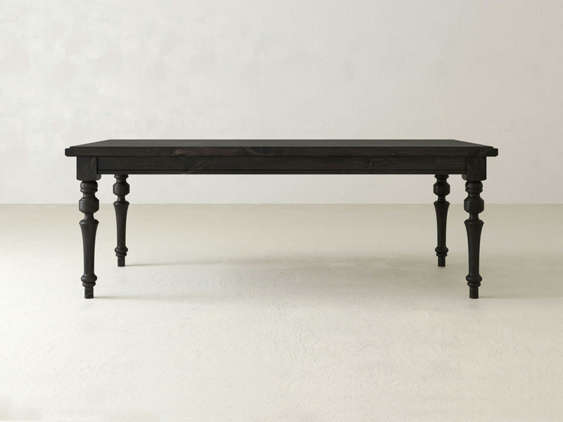 The Allyson Dining Table - Charred Ember by James+James, featuring intricately carved legs, stands against a plain, light-colored background. The table has a rectangular shape with a smooth, polished surface that highlights its minimalist yet elegant design.