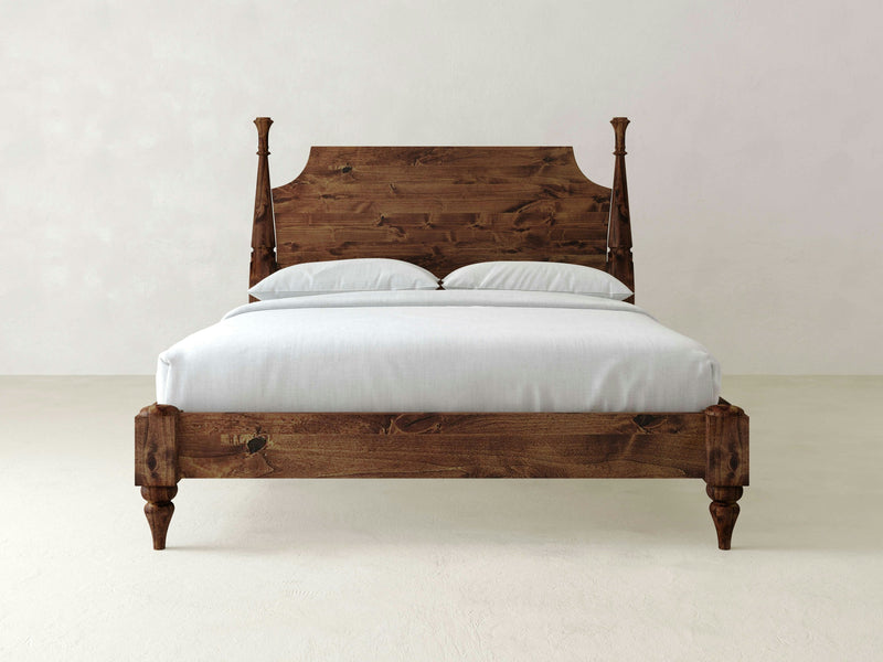 A Stella Bed - Tuscany by James+James is showcased against a plain white background. The wooden bed frame features a tall, intricately carved headboard and four sturdy legs. It is dressed in white bedding with two matching white pillows, highlighting its rustic design with a dark wood finish.