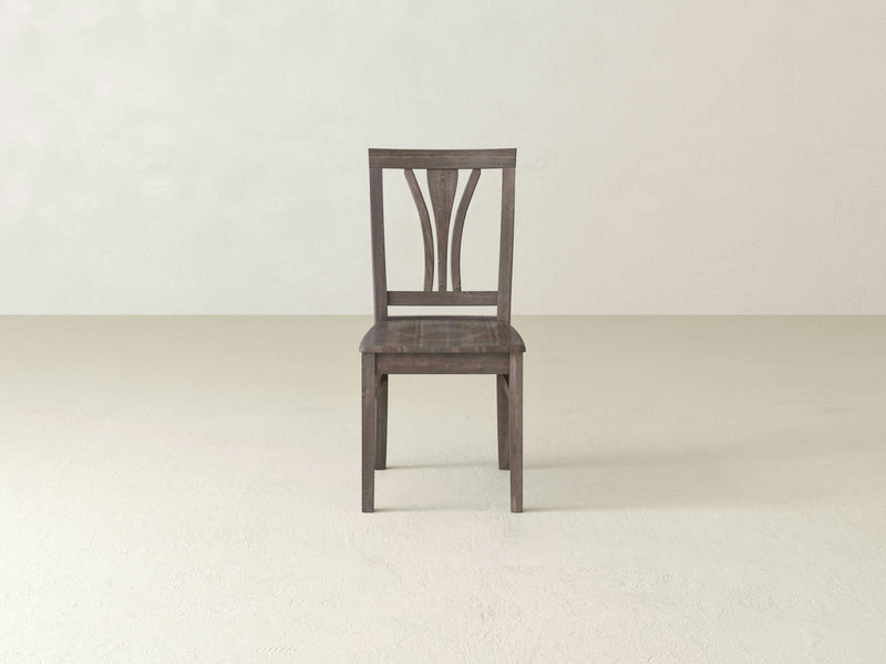 The Brooklyn Dining Chair - Barn Wood by James+James with its minimalist design rests on a light-colored floor. The chair features a solid backrest adorned with vertical and curved slats forming an intricate pattern. Its overall color is a rich, dark brown or greyish tone, set against a plain and minimalistic background.