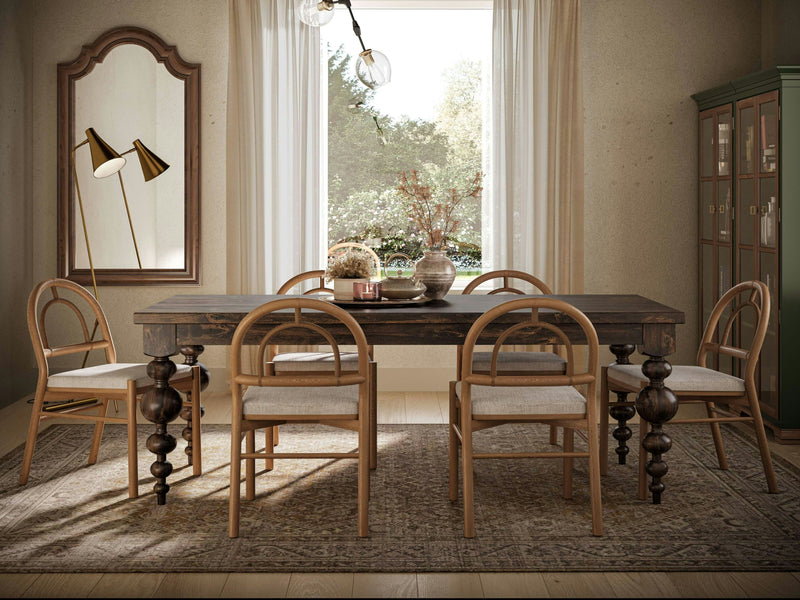 A dining room featuring the Olivia Expandable Dining Table in a rich Tobacco finish from James+James, accompanied by six chairs, all set on a patterned rug. To the left stands a large mirror and a brass floor lamp, while on the right there is a green cabinet. Natural light streams through sheer curtains covering a window, revealing an outdoor view filled with greenery.