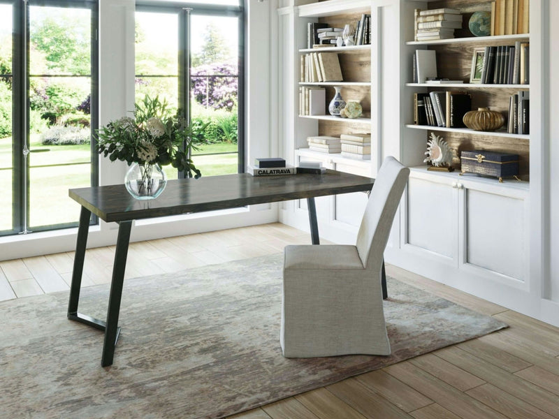 A modern home office featuring the Arkwright Desk in Deep Grey from James+James, paired with a padded chair. The desk is adorned with a vase of fresh flowers. Large windows offer a view of a lush garden. Bookshelves filled with decor and books flank the desk, set against white-paneled walls.