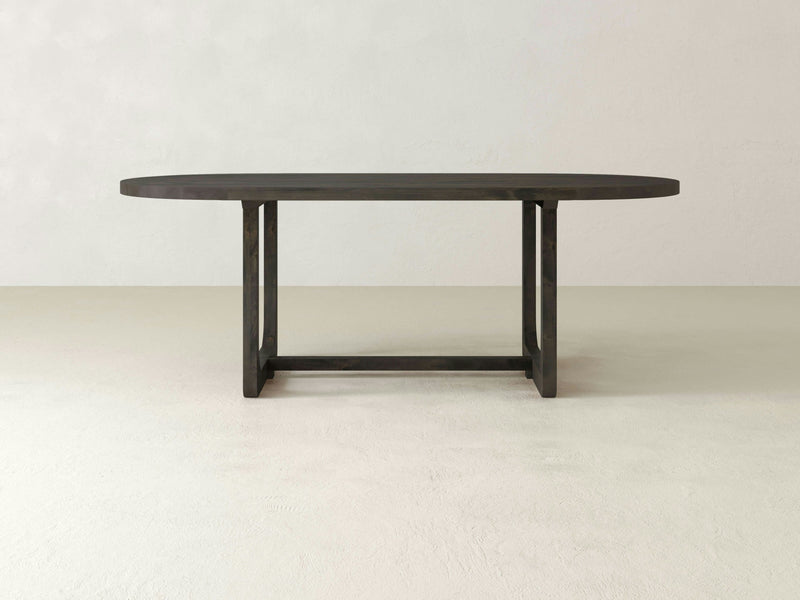 The Emersen Oval Dining Table by James+James, featuring a deep grey finish, stands on a light-colored, textured floor. The table boasts clean lines and a modern trestle base design with two vertical supports connected by horizontal beams. The background is plain and light-colored.