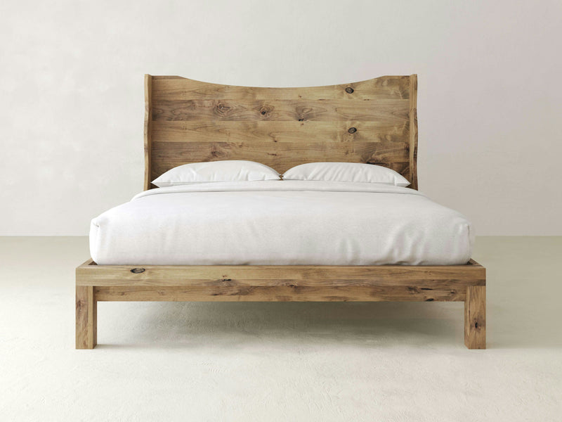 The James+James Harlow Bed exudes natural beauty with its solid wooden frame and headboard in a natural wood finish. This elegant wingback design, adorned with white bedding and two pillows, sits against a light-colored wall in a minimalist room, highlighting the simplicity and quality materials.