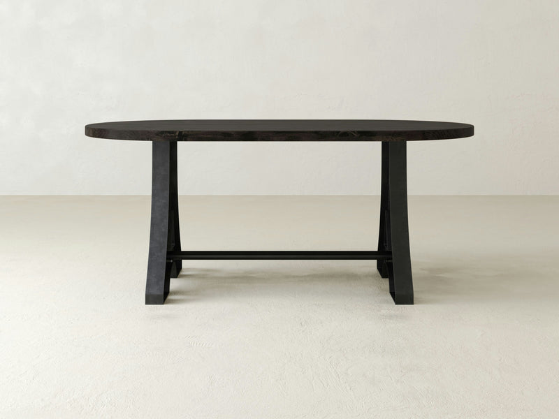 The Wishbone Oval Dining Table - Charred Ember by James+James is a minimalist, oval-shaped table with a sleek design. It features two sturdy, angled legs connected by a horizontal support beam. The background showcases a plain, light-colored wall and floor, creating a simple and elegant setting.