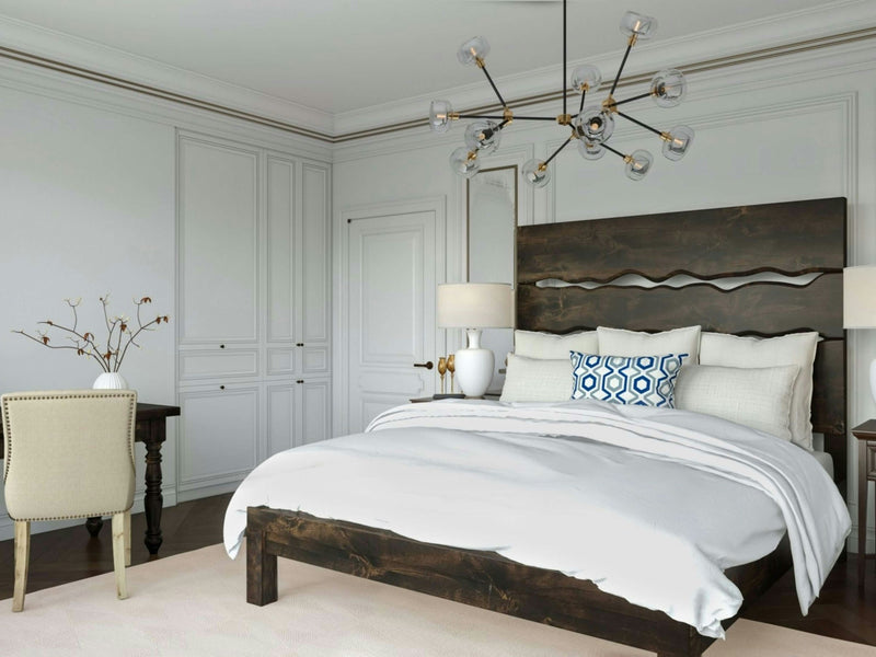 A stylish bedroom features a James+James Live Edge Bed in Charred Ember with white linens and a blue and white decorative pillow. A unique chandelier hangs from the ceiling. A beige chair and desk sit in the corner, and the walls are adorned with minimalist white paneling and trim.