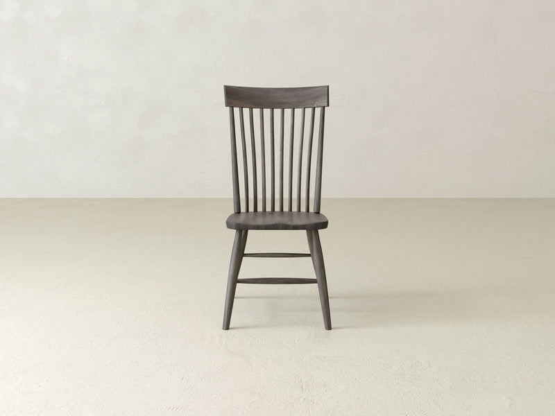 An Eleanor Side Dining Chair in deep grey from James+James stands alone on a plain, light-colored floor against a light-colored wall. The chair has vertical slats on the backrest and embodies a simple, classic design.