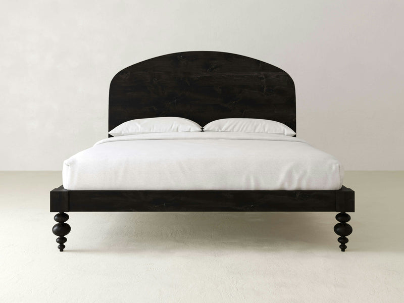 The Eden Bed by James+James is a minimalist dark wood bed frame with a curved headboard, complemented by two white pillows and a neatly made white bedspread. Crafted from solid hardwood, the bed stands on four turned legs against a light-colored floor and plain, light-colored wall, making it an ideal choice for any bedroom decor.