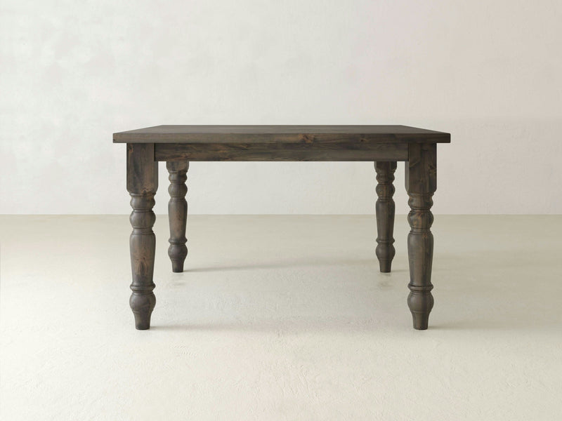 The **Baluster Square Dining Table - Deep Grey** by **James+James** stands on four intricately carved legs. The table is positioned against a light-colored, plain wall and floor, highlighting its rustic texture and craftsmanship.