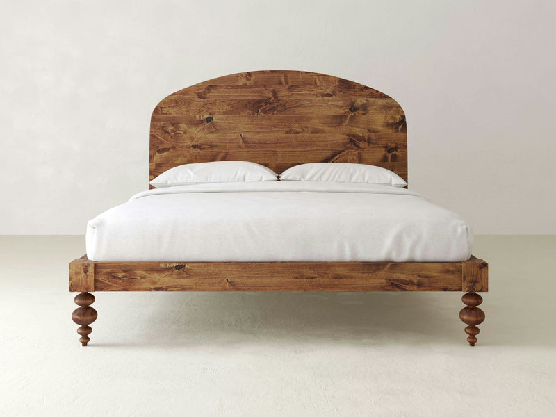 The Eden Bed - Tuscany by James+James is shown, featuring a curved headboard and turned legs. Made from rich, polished wood, this wooden bed frame exhibits a rustic aesthetic. It is elegantly dressed with white bedding, including pillows and a duvet.