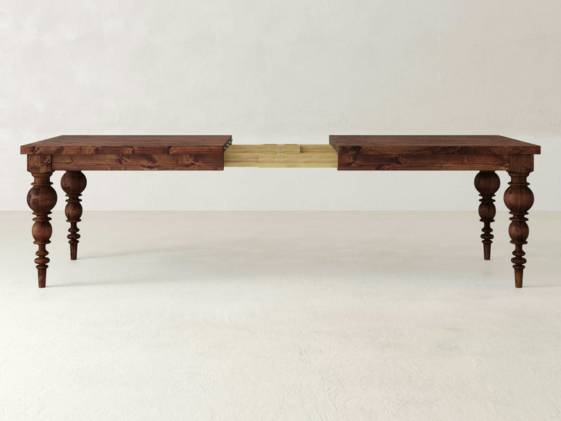 The Bailey Expandable Dining Table - Tuscany by James+James features ornate, carved legs and showcases a rich, dark finish. The table is displayed in its extended position, revealing a visible gap where an extension leaf would be placed. The legs are intricately detailed with turned designs.