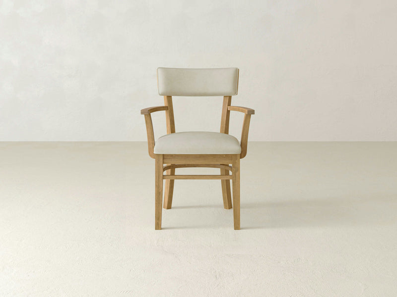 The Everly Arm Dining Chair in Harvest Wheat by James+James features a wooden frame and a beige cushioned seat and backrest. This chair includes armrests and is placed in a minimalist setting with a light-colored floor and an off-white textured wall as the backdrop.