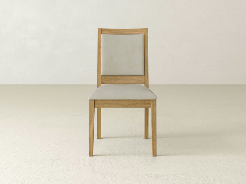 A Gina Side Dining Chair - Harvest Wheat by James+James, featuring a minimalist wooden design and a light grey cushioned seat and backrest, placed against a plain off-white wall and floor background. The chair boasts a clean, modern aesthetic with straight legs and a rectangular back frame.