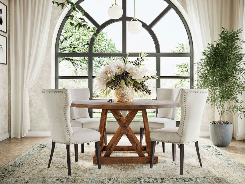 An elegant dining room features the Julia Round Dining Table - Tuscany by James+James, complete with a vase of large white flowers. Four beige upholstered chairs surround the table. The room is adorned with a large arched window, cream curtains, a potted plant, and a light-colored patterned rug.