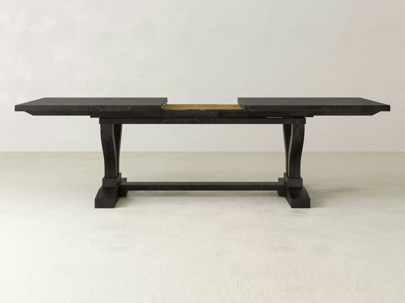 The Vera Expandable Dining Table - Charred Ember by James+James is depicted. The wooden table, with its dark finish, features an extending design and has a central leaf removed, creating a gap in the center. The table sits on a sturdy rectangular base with two pedestal legs. The background is plain and light-colored.