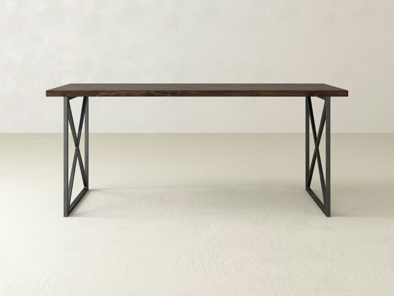 The Luca Desk - Tobacco by James+James features a rectangular wooden top in a dark brown finish and black metal legs arranged in a crossed pattern. This modern and minimalist design provides a sturdy and stylish workspace, set against a plain white background.