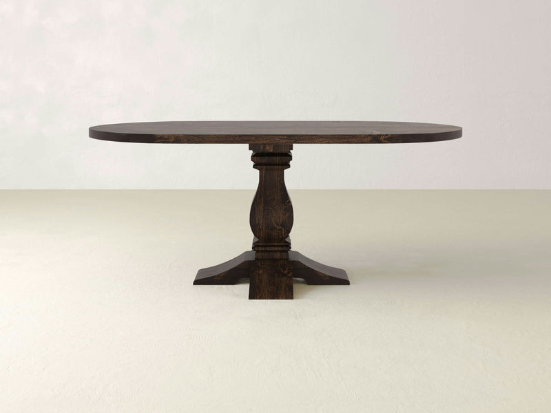 The Heirloom Oval Dining Table - Tobacco by James+James stands on a light-colored surface against a plain, off-white background. This dark wooden table features a single pedestal base, showcasing its classic, elegant design with a smooth finish and a sturdy, ornamental central support.