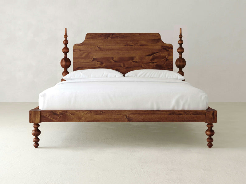 In a minimalist grand bedroom, a wooden Opal Bed from James+James stands prominently with its tall, carved headboard and four decorative turned posts, each adorned with finials. The bed features a white mattress and two white pillows, all supported by an elegant frame elevated by turned wooden legs.