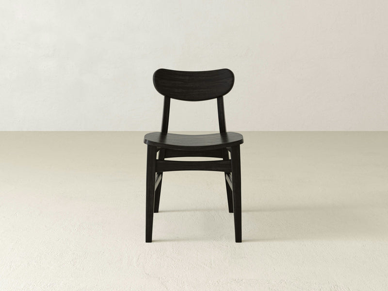 The Lynn Dining Chair - Charred Ember by James+James features a minimalist design with a modern and sleek appearance. It has a curved backrest and four sturdy legs, crafted from black wood. Positioned on a light, textured floor against a plain neutral wall, this chair exudes simplicity and elegance.