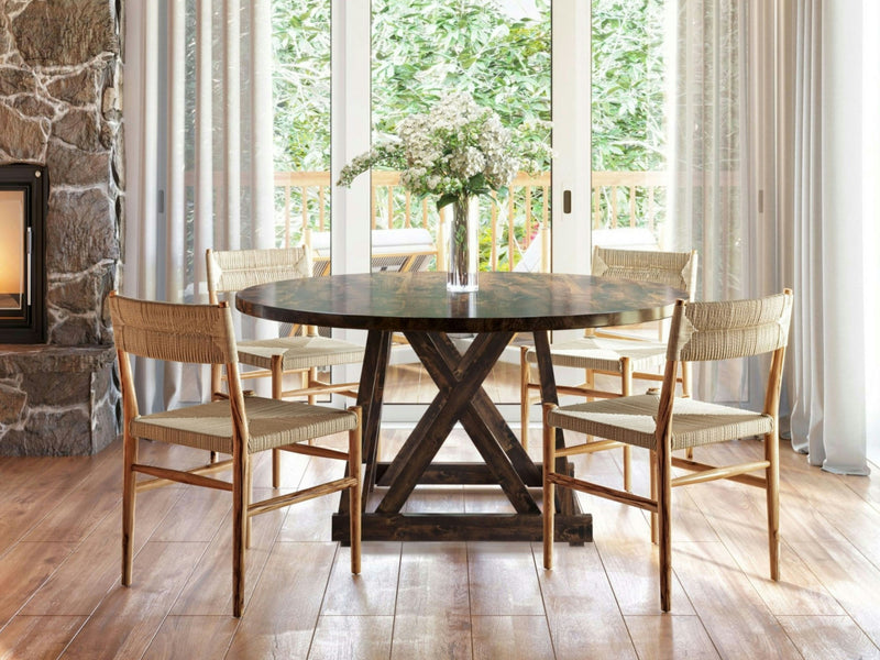 A dining area features the Julia Round Dining Table in Tobacco by James+James, surrounded by four wooden chairs with woven seats. In the center of the table, there's a vase with white flowers. The background includes large glass sliding doors, sheer curtains, and a stone accent wall.