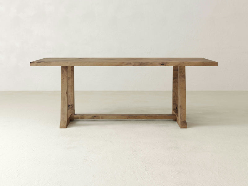 A minimalist, rectangular wooden table by James+James, known as the Cora Dining Table in Harvest Wheat finish, with a flat top and sturdy legs is displayed in a white, empty room. Its design is simple and functional, featuring natural wood aesthetics. The legs of the table are connected by a horizontal support bar near the floor.