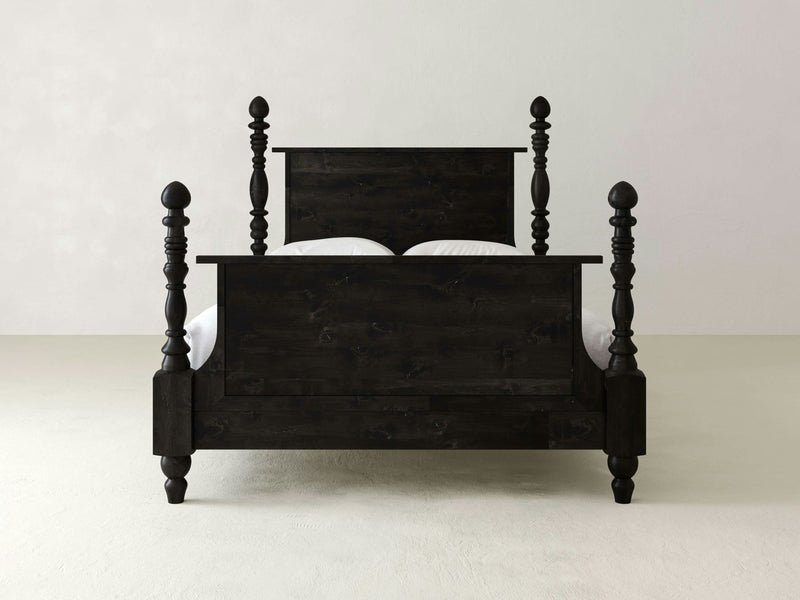 The Catherine Bed - Charred Ember by James+James, showcasing a dark wooden finish with a high headboard and footboard, is positioned against a plain, light-colored wall. The bed features turned posts with finials at each corner and boasts a traditional design with visible wood grain.