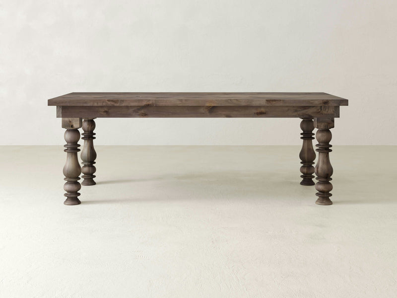 The Annli Dining Table by James+James is a rustic piece with a rectangular top and sturdy legs that showcase intricate carvings. Set against a plain, light-colored background, the table has a distressed finish that enhances its rustic charm.