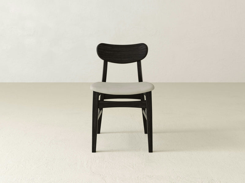 In the center of a light-colored, empty room with plain-textured walls and floors, the Lynn Upholstered Dining Chair - Charred Ember by James+James is placed. This minimalist chair features a dark wooden backrest and legs paired with a cushioned light gray seat, embodying a modern and simple design that emphasizes clean lines and simplicity.