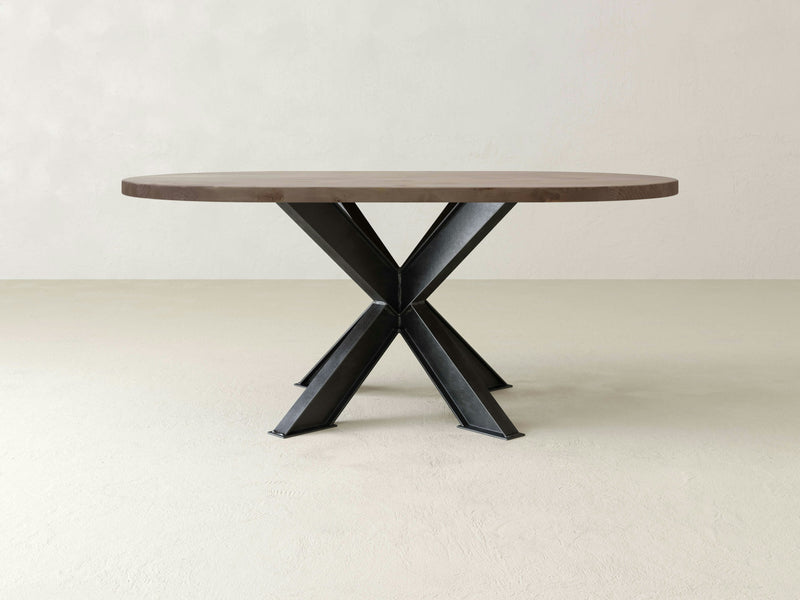 The Shiloh Oval Dining Table - Barn Wood by James+James stands in the center of a minimalist room. The table features a thick top with a modern design and a unique black metal base with four legs converging at the center and spreading out at angles, creating an X-shaped support structure.