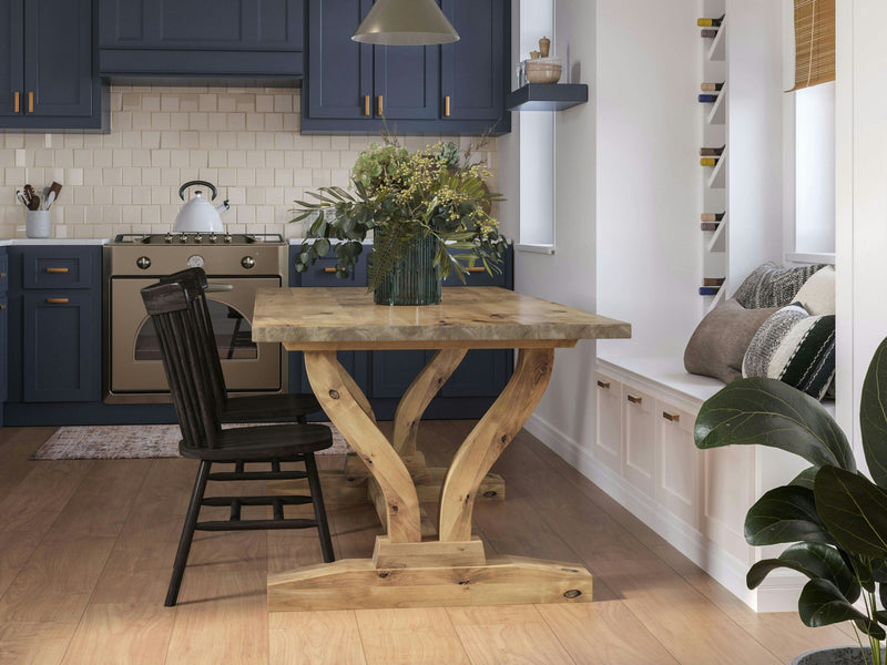 A cozy kitchen featuring blue cabinets, a James+James Vera Dining Table in Harvest Wheat, and a black chair. The backsplash is white tile, accompanied by a beige stove. There is a built-in bench with cushions and decorative shelves on one side. A large vase with greenery serves as the centerpiece on the table.
