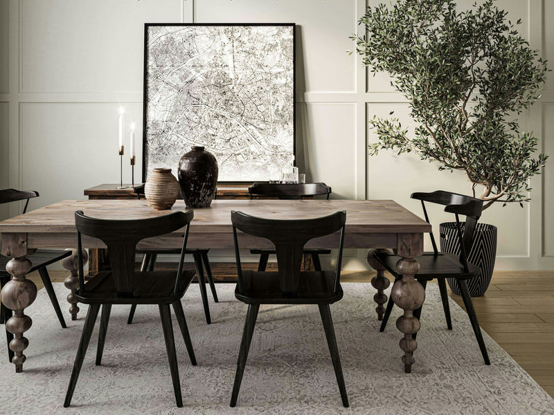 A stylish dining room features the Olivia Dining Table in Barn Wood from James+James, surrounded by four dark chairs. The table is adorned with various decorative items, including a large dark vase, smaller vases, and tall candles. To the right stands a tall leafy plant, and textured artwork graces the wall behind.