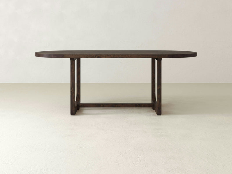 An Emersen Oval Dining Table - Tobacco by James+James with a wooden top and sturdy, rectangular legs is placed in a minimalist room with neutral-colored walls and floors.