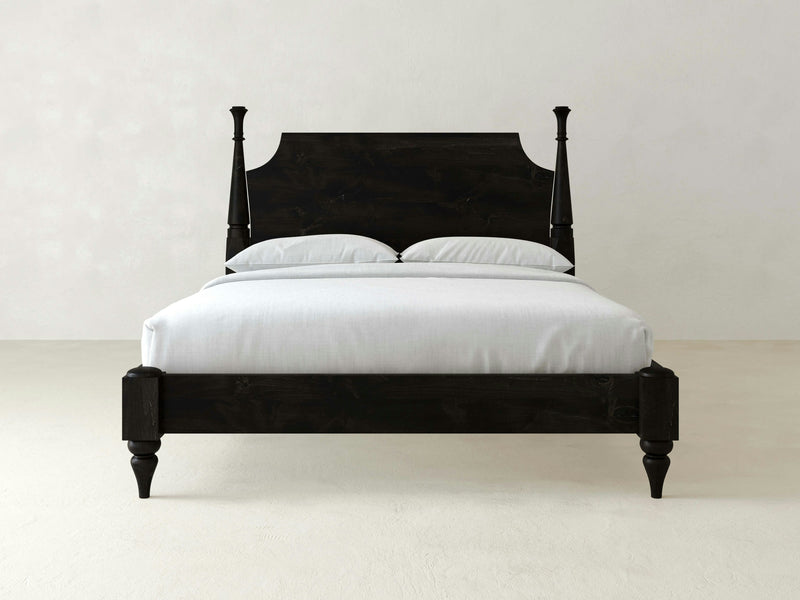 The Stella Bed - Charred Ember by James+James, a sleek, minimalist black wooden four-poster bed, is showcased against a plain light background. The bed is impeccably made with white linens. Its tall headboard features curved details, and each corner of the frame boasts turned wooden posts.